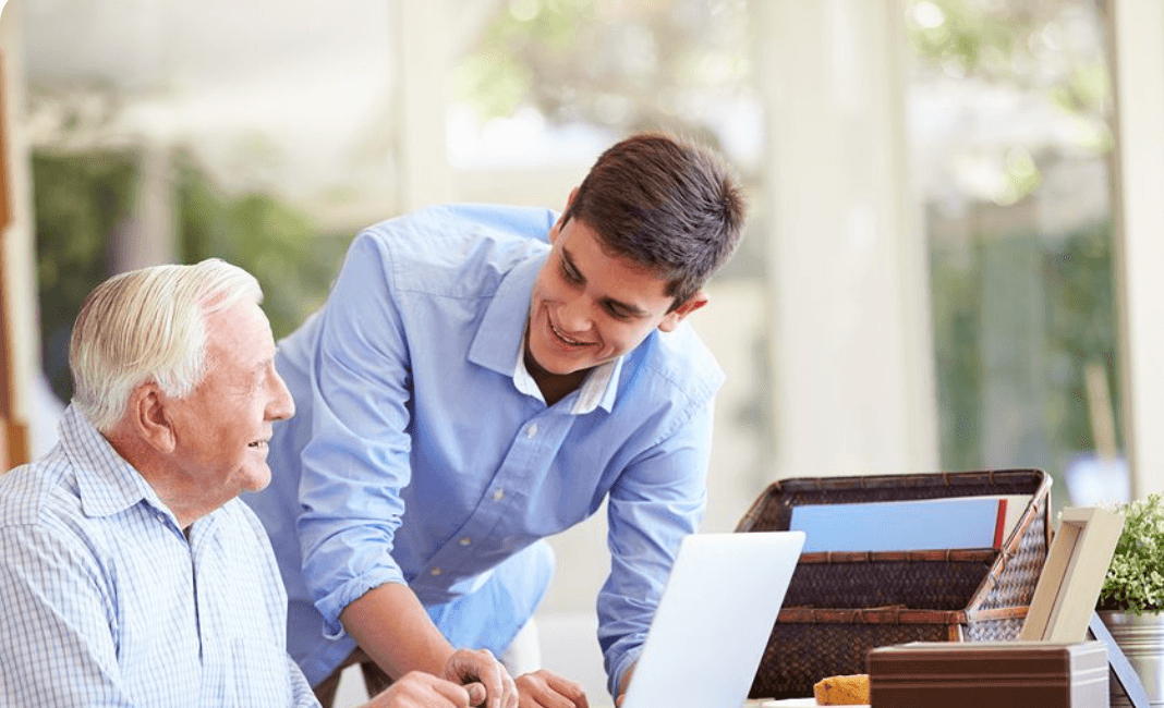 A Tech Peace technician leaning in beside an older man at his dining table, both smiling as they look at his laptop together