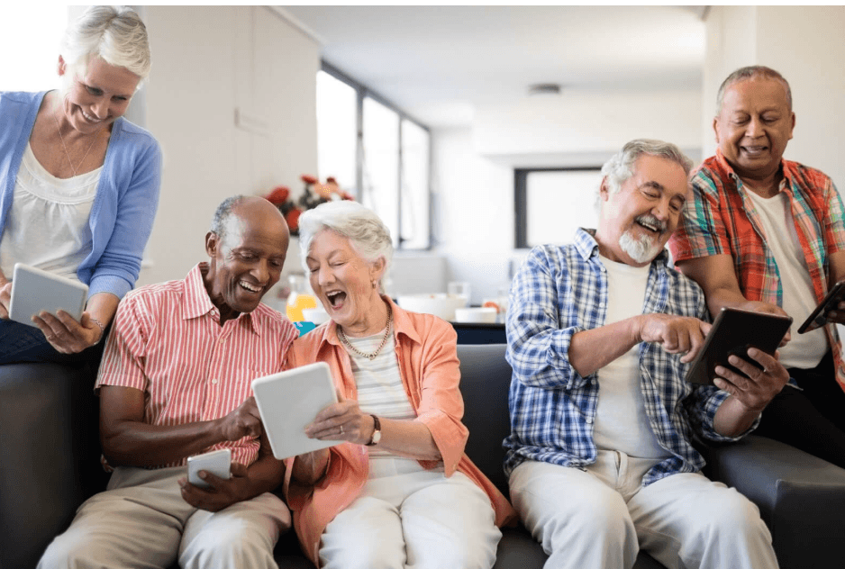 A group of older adults sitting together on a sofa with tablets, laughing at something on screen