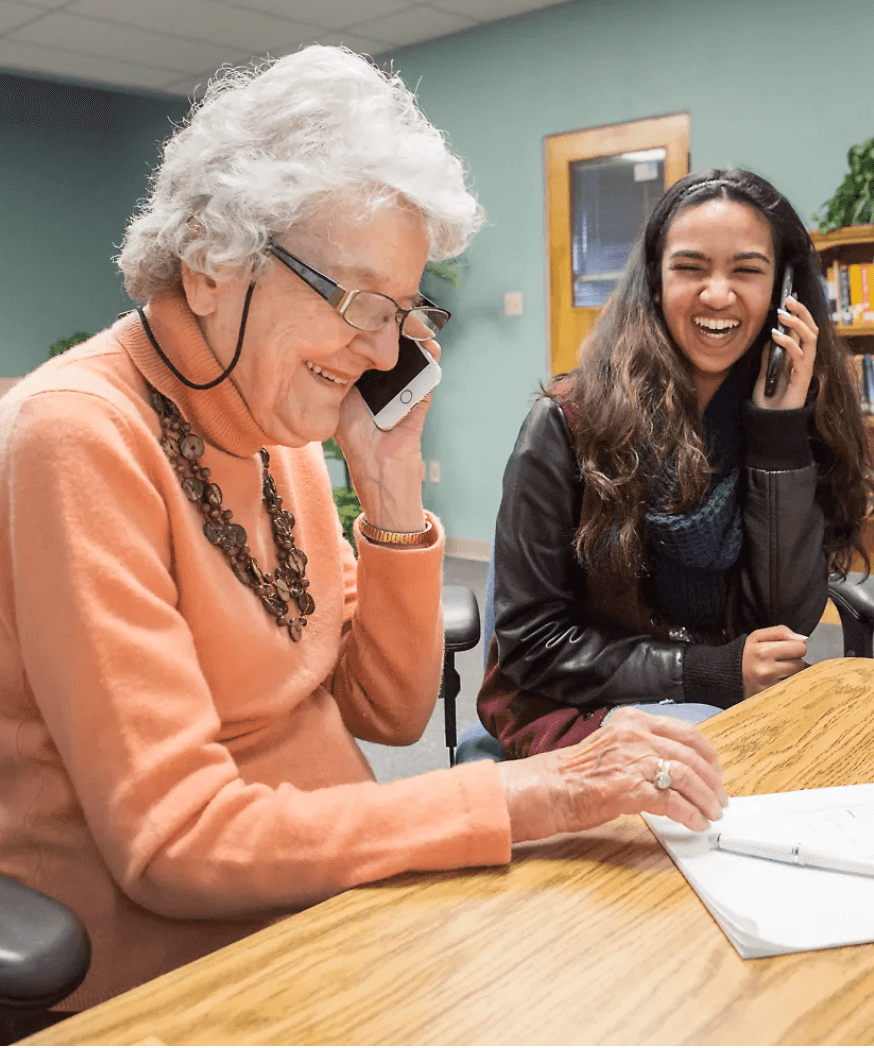 An older woman and a young woman sitting side by side at a table, both on their phones and laughing together