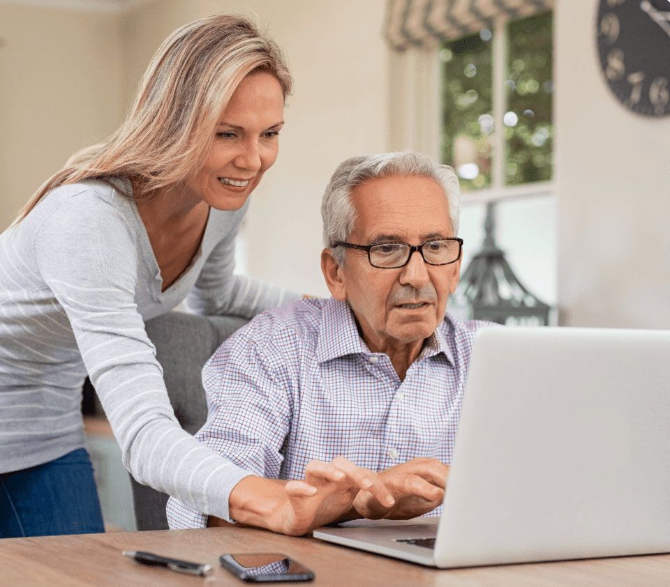An adult daughter leaning over her father at the kitchen table, pointing something out on his laptop