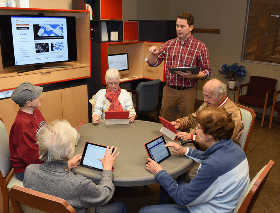 An instructor standing at a table leading six older adults through a tablet class in a community room